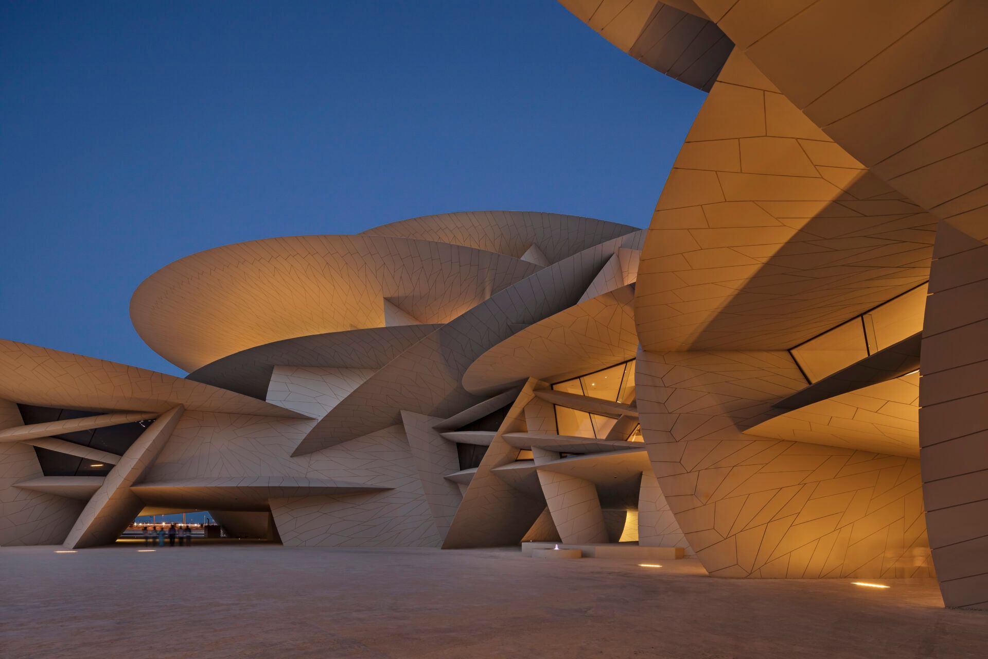 The National Museum of Qatar facade,  illuminated from below at dusk against a dark blue sky.