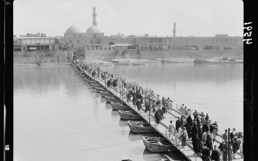 A vintage black and white photograph of crowds walking up a long bridge or pier, in historic Baghdad.