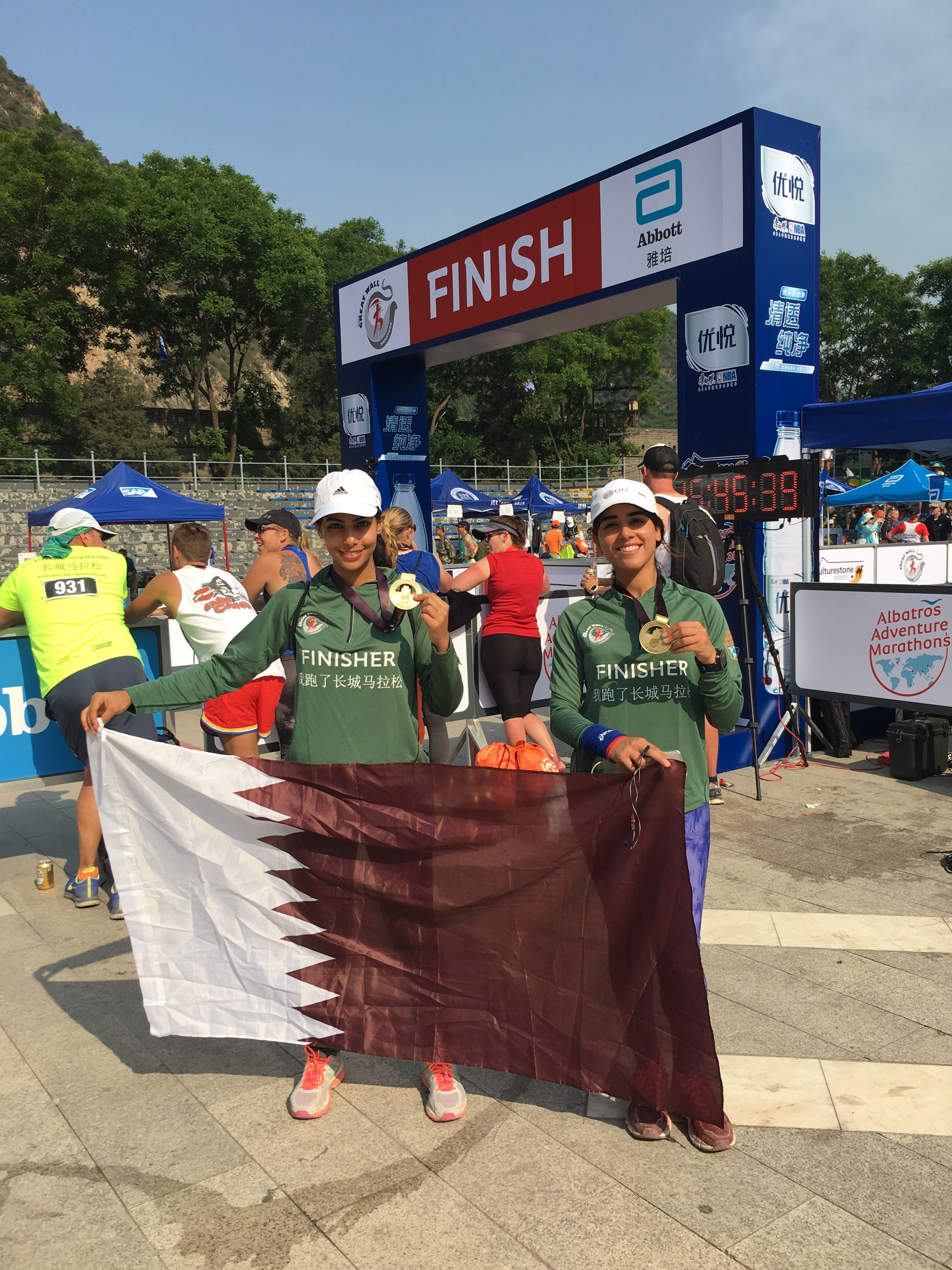 2 Qatari women in green sweatshirts hold a Qatari flag and gold medals at the finish line of a marathon.