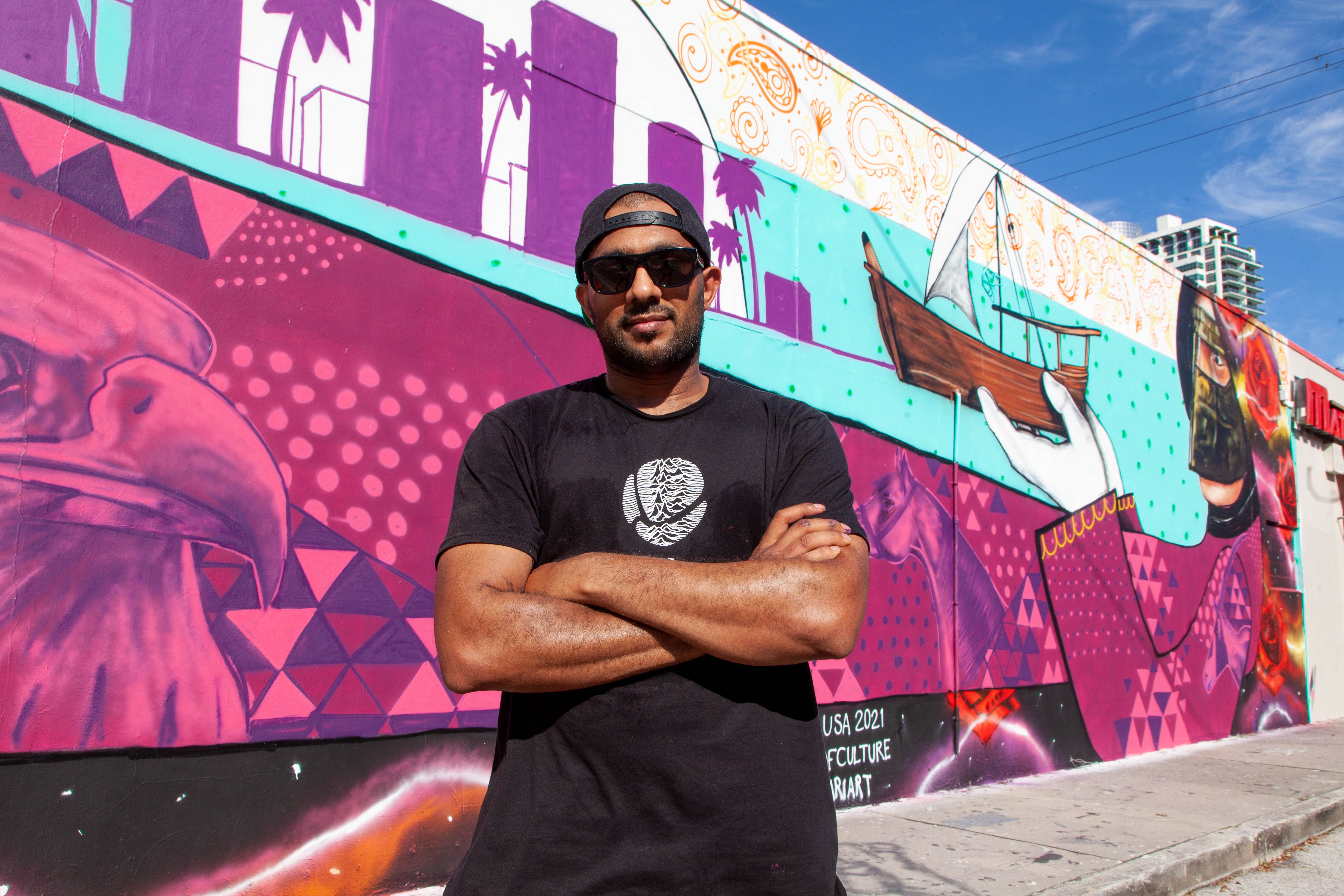 Man wearing a black tshirt and baseball cap stands with arms crossed in front of a colourful JEDARIART mural in Miami.
