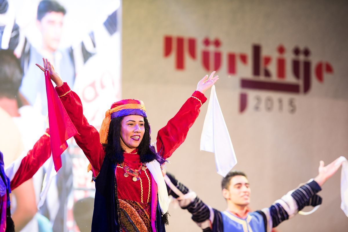 Woman wearing a traditional Turkish costume on stage with her arms up in the air holding a fabric scarf in each hand.