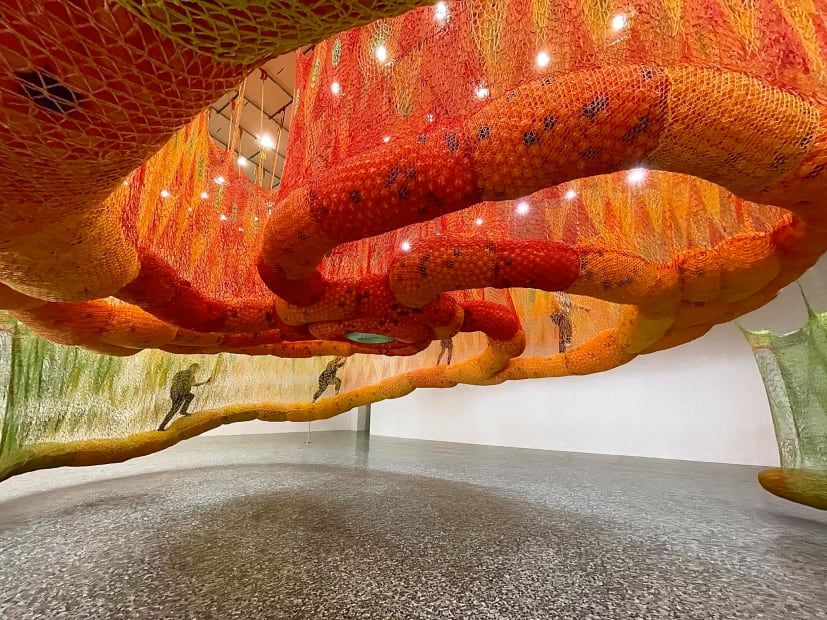 Large orange art installation by Ernesto Neto of fabric walkways hung from the ceiling in a gallery space.